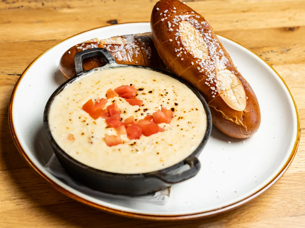 A cast iron dish of warm dip topped with diced tomato, with soft pretzel bread